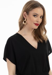 Schwarze Bluse mit V-Ausschnitt und kurzen Ärmeln. Das Model trägt goldene, texturierte Creolen. Lange, wellige braune Haare umrahmen das Gesicht.
