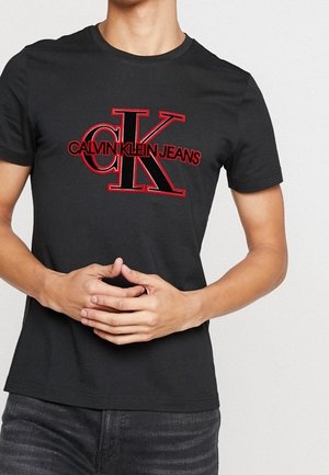 Person trägt ein schwarzes T-Shirt mit großem roten und schwarzen "CK Calvin Klein Jeans" Logo, Hände locker vor dem Körper verschränkt.