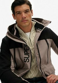 Kleurblokkering jack in beige en zwart, met een rits aan de voorkant, hoge kraag en geborduurd logo. Gelaagd over een crèmekleurige kabelgebreide trui.