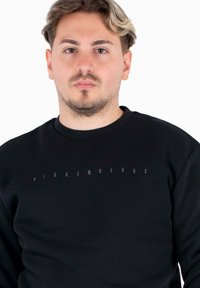 Giovane uomo con capelli corti e una felpa nera Bikkembergs, di fronte a uno sfondo bianco semplice.