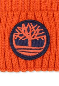 Beanie orange tricoté avec une texture côtelée, doté d'un patch rond bleu avec un motif d'arbre brodé et un détail de ligne horizontale.