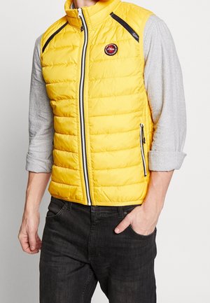 Homme portant un gilet matelassé jaune vif avec des détails de fermeture éclair noirs par-dessus une chemise à manches longues gris clair et un jean foncé, main dans la poche.