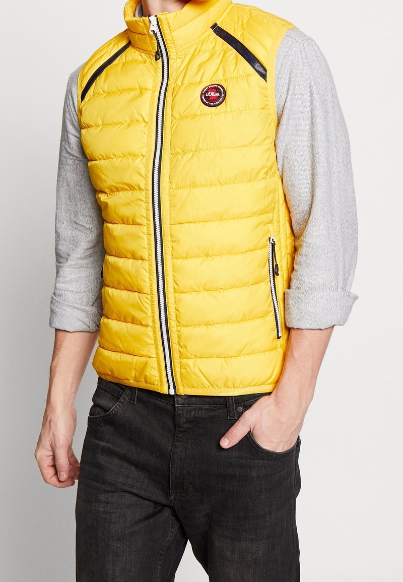 Homme portant un gilet matelassé jaune vif avec des détails de fermeture éclair noirs par-dessus une chemise à manches longues gris clair et un jean foncé, main dans la poche.