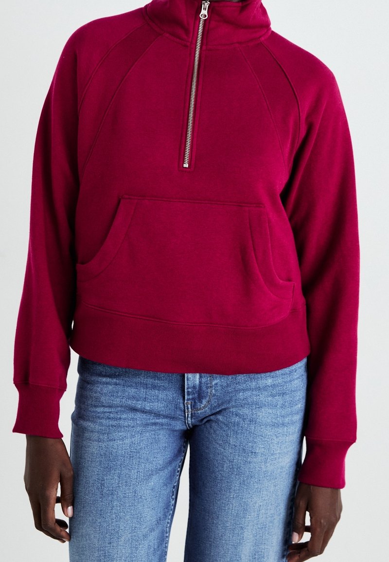 Personne portant un sweat-shirt rouge framboise avec fermeture éclair quart de zip et une poche avant ainsi qu'un jean bleu, vue du cou jusqu'aux hanches.