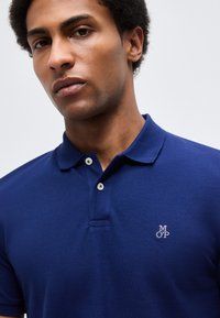 Νεαρός άνδρας που φοράει στενή μπλε πόλο μπλούζα με γιακά και δύο κουμπιά, με μικρό λευκό λογότυπο "MOP" στο στήθος.