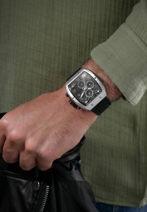 Vierkante zilveren horloge met een zwarte rubberen band, doorzichtige wijzerplaat die het binnenwerk onthult, en meerdere wijzerplaten, gedragen aan een hand die een zwarte tas vasthoudt.