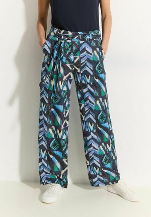 Osoba ubrana w spodnie z szerokimi nogawkami o niebiesko-zielono-czarnych geometrycznych wzorach, z dopasowanym paskiem, czarną bluzkę i białe trampki.