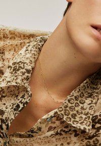 Un collar de cadena dorada descansa sobre la clavícula, acompañado de una camisa de leopardo de color marrón claro con un cuello clásico y un botón azul.