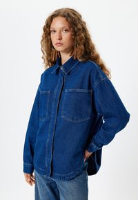 Camisa de mezclilla azul con un corte relajado, que presenta dos bolsillos en el pecho, un cierre frontal con botones y mangas largas. La textura es suave y resistente.