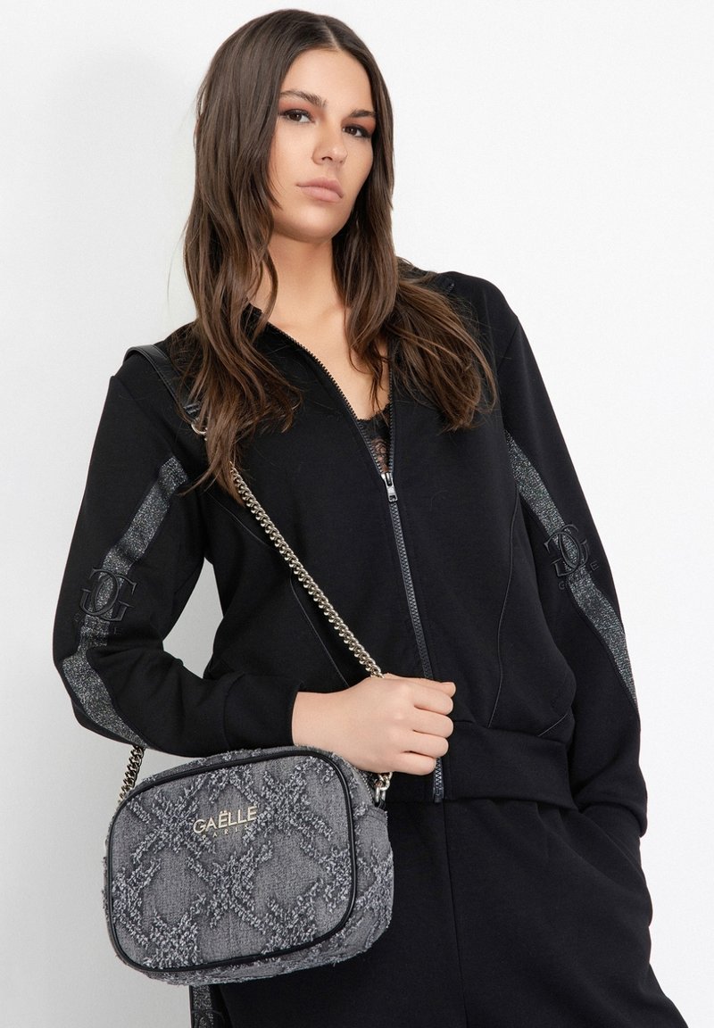 Giacca nera con zip e dettagli argentati, abbinata a una borsa grigia texturizzata con motivo decorativo e tracolla a catena.