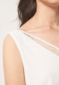 White top featuring a wide, asymmetrical neckline with lace detail along the shoulder, textured fabric, and a fitted design.