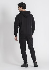 Felpa nera con cappuccio, maniche lunghe, polsini e orlo a coste. Indossata con jeans scuri e sneakers nere, vista da dietro.