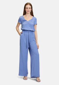 Combinaison bleu clair avec des manches courtes, un col en V et une taille à cordon de serrage ; présente des pantalons à jambes larges et une texture de tissu lisse.