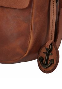 Braune Lederhandtasche mit glatter Textur, ausgestattet mit einem Frontklappe und einem runden Etikett mit einem Ankeremblem. Sichtbare Nähte.