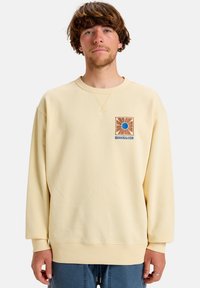 Cream-colored sweatshirt with a round neckline, ribbed cuffs, and a square embroidered patch featuring a sun design and logo on the chest.
