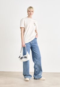 Šviesiai smėlinis medvilninis marškinėlis su spalvinga grafika ant krūtinės, derinamas su šviesiai mėlynomis plačiomis kelnėmis ir baltu rankiniu krepšiu su grandinės detalėmis.