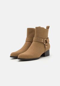 Botas de tobillo de ante color tan con tacón cuadrado, que presentan una correa decorativa con tachuelas redondas y una lengüeta en la parte posterior para facilitar su uso.