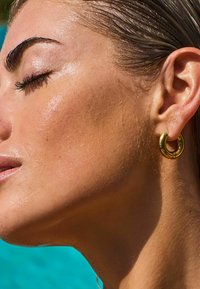 Boucle d'oreille en or sur l'oreille d'un modèle, avec un design élégant et arrondi. La peau paraît scintillante, sur fond turquoise.
