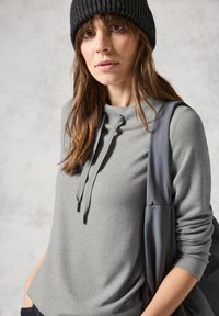 Grå tröja med hög hals och snörning; ribbad textur. Bärs med en mörk mössa och en grå axelväska. Neutral bakgrund.