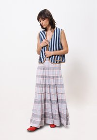 Gestreiftes blau-weißes Weste mit sandfarbenen Akzenten, über einem fließenden, gestuften Maxirock mit einem pastellfarbigen Karomuster, geschichtet. Rote Schuhe vervollständigen den Look.
