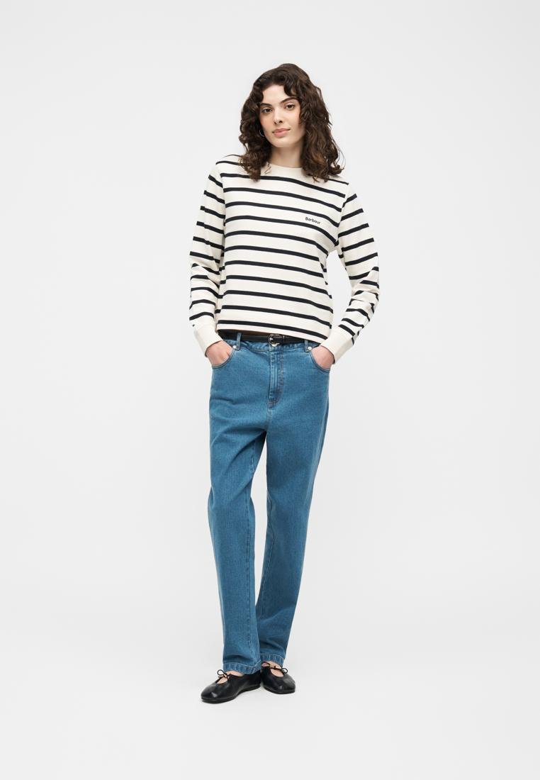 Femme aux cheveux bouclés portant un t-shirt à manches longues blanc et noir à rayures, un jean bleu, une ceinture noire et des ballerines noires, debout les mains dans les poches.