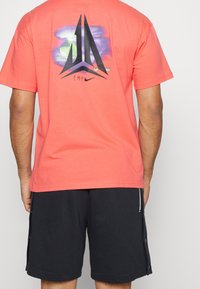Coral cotton t-shirt with black logo design and gradient background. Paired with dark shorts featuring side buttons and zipper details.