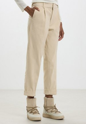 Person trägt beige Cordhosen mit hochgekrempelten Bündchen und beige hohe Schnür-Freizeitschuhe, steht mit einer Hand in der Tasche.