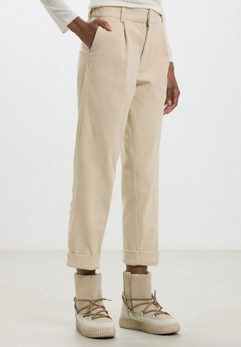 Personne portant un pantalon en velours côtelé beige avec des revers roulés et des bottines montantes beiges lacées, debout avec une main dans la poche.