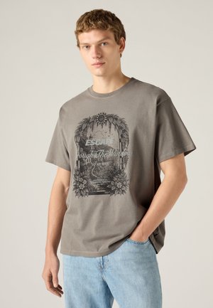 Jeune homme aux cheveux courts et bouclés portant un t-shirt gris à motif et un jean bleu clair, une main dans la poche, sur fond uni.