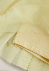 Kollane tüllikate, mis on kihina pehme kollase riide kohal, kaunistatud õrna lillemustriga tikanditega ja korralike õmblustega äärte ümber.