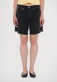 Shorts in denim neri con orlo sfrangiato, due tasche frontali e dettagli in metallo argentato. Indossati con una maglietta gialla e scarpe nere flat.