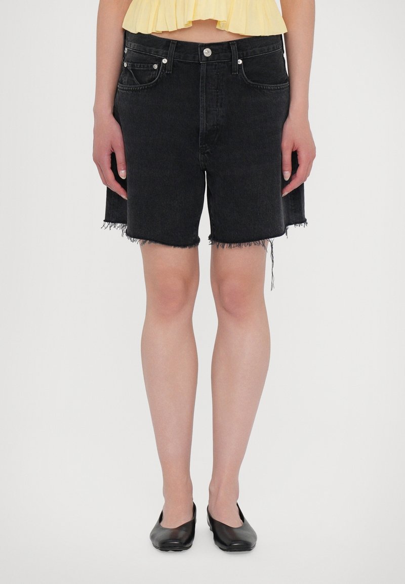 Shorts in denim neri con orlo sfrangiato, due tasche frontali e dettagli in metallo argentato. Indossati con una maglietta gialla e scarpe nere flat.