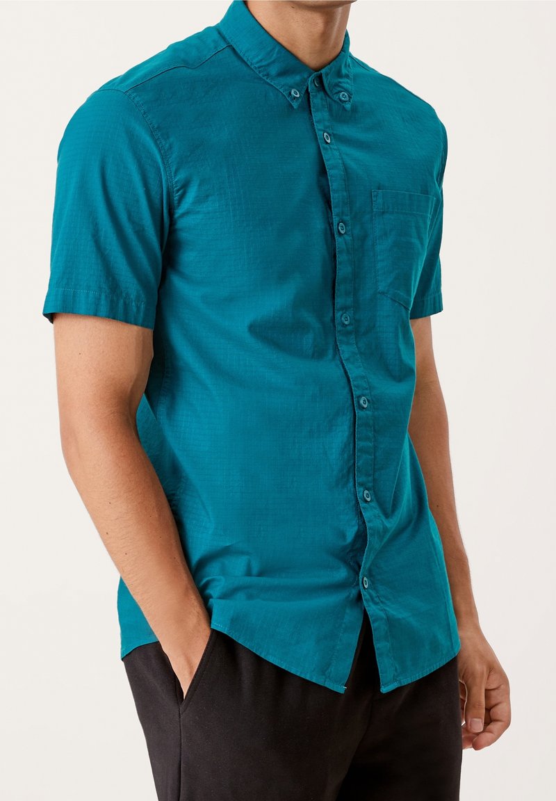 Camicia a maniche corte di colore teal con taschino sul petto; il tessuto leggero presenta una sottile trama e un colletto.