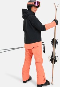 Echipament de schi format dintr-o jachetă neagră cu accente portocalii și pantaloni portocalii asortați, echipat cu o cască și bețe de schi.
