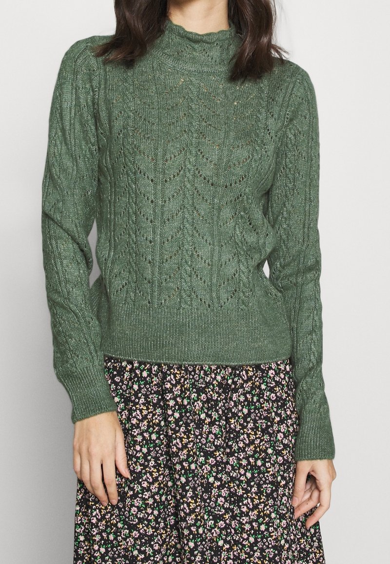 Pull en tricot vert avec un col montant, présentant un motif texturé en forme de feuille et un ourlet côtelé, associé à une jupe à imprimé floral.