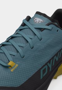 Μπλε αθλητικό παπούτσι με επάνω μέρος από πλέγμα, κορδόνια τιρκουάζ, κίτρινο θηλάκι κορδονιών, μαύρη σόλα και λογότυπο στη γλώσσα και στην πλευρά που γράφει "DYNA".