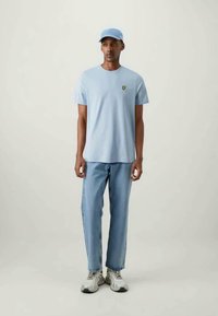 Tricou din bumbac de culoare albastru deschis, cu mâneci scurte și un logo verde, asortat cu blugi de culoare albastru deschis și o șapcă albastră corespunzătoare, purtând adidași bej.