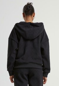 Person mit einem Haarknoten und Ohrringen, die einen schwarzen strukturierten Hoodie und passende Hosen trägt und von hinten gesehen wird, mit einem Halbmond-Tattoo am Nacken.
