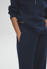 Une personne portant un haut à manches longues côtelé bleu marine et un pantalon assorti, les mains partiellement dans les poches, visible du milieu du torse aux cuisses.
