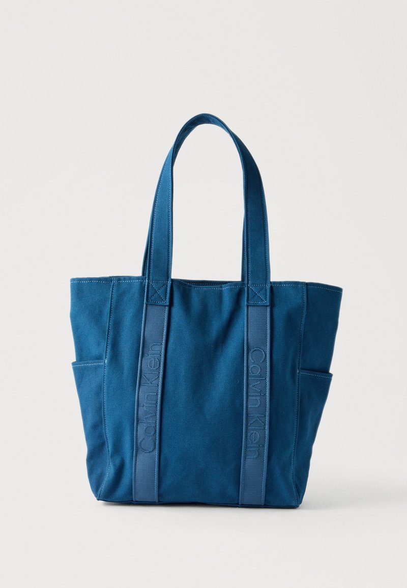 Sac cabas en toile bleue Calvin Klein avec deux poignées et poches latérales, sur un fond clair uni.