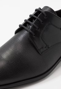 Chaussure habillée en cuir noir avec texture perforée, panneaux lisses, six œillets et lacets plats, dotée d'un bout arrondi et d'une semelle noire.