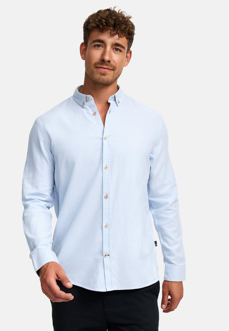 Giovane uomo con capelli ricci che indossa una camicia azzurra con bottoni e pantaloni neri, in piedi davanti a uno sfondo bianco semplice.