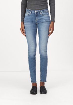 Kvinde iført lyseblå skinny jeans, sorte loafers og en grå langærmet top står mod en hvid baggrund.