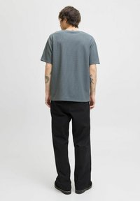 Person set fra bagfra, iført en løs grå t-shirt og sorte bukser, med synlige tatoveringer på armene, sat mod en enkel hvid baggrund.