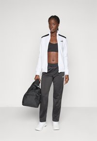 Vitt zip-up-jacka med svarta detaljer, svart sport-bh, grå träningsbyxor och vita sneakers. Håller en svart axelväska.