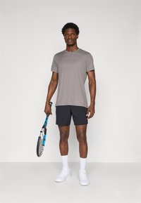Grå atletisk t-shirt, sorte shorts og hvide sneakers. Holder en tennisketsjer med blå og sorte accenter. Enkel, funktionel design.