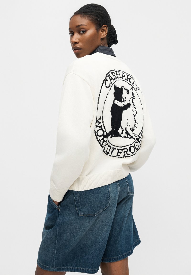 Donna che indossa un maglione bianco con grafica di gatto bianco e nero e testo "CARHARTT WORK IN PROGRESS", abbinato a pantaloncini di jeans blu.