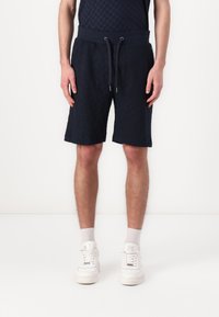Pantalones cortos de algodón azul marino con una cinturilla elástica, cierre de cordón y un patrón texturizado. Combinados con zapatillas blancas y calcetines por el tobillo.