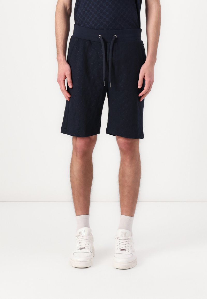 Pantalones cortos de algodón azul marino con una cinturilla elástica, cierre de cordón y un patrón texturizado. Combinados con zapatillas blancas y calcetines por el tobillo.