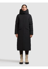 Must pikk sulejope kõrge kraega, lukuga, kahe esitasku ja vooluva disainiga. Kanditud mustade kitsaste retuuside ja beežide saapadega.